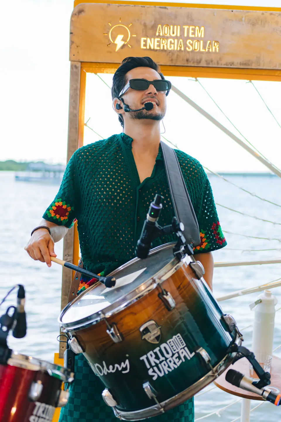 Hegon Nunes de óculos escuros e camisa de crochê verde tocando a zabumba com a logo do trio