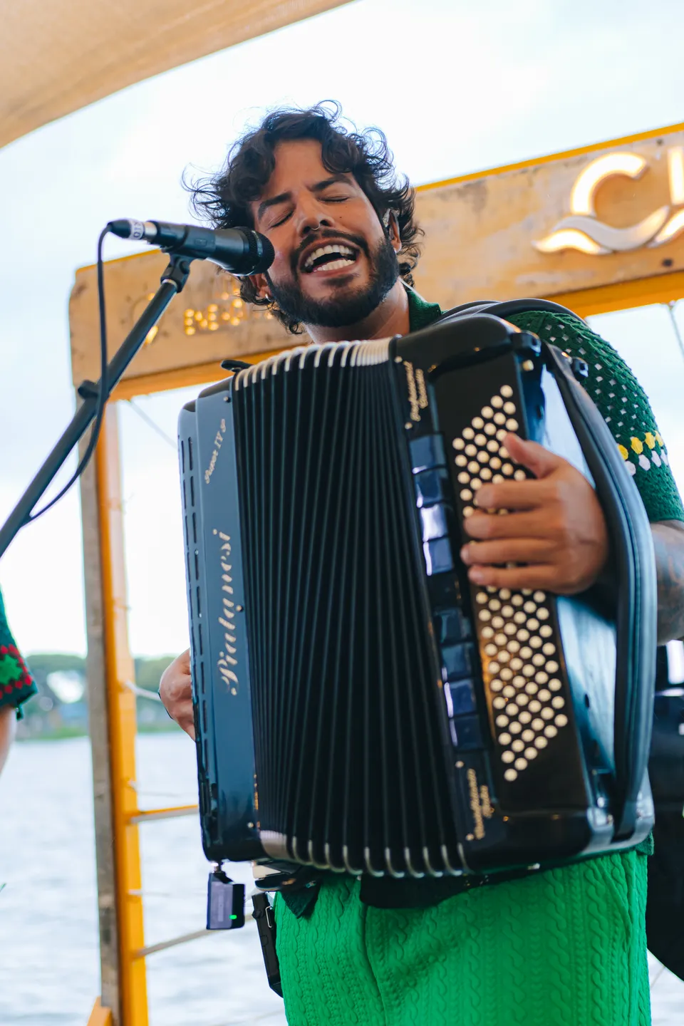 Lucas Menezis cantando ao microfone enquanto toca sanfona num palco à beira do rio
