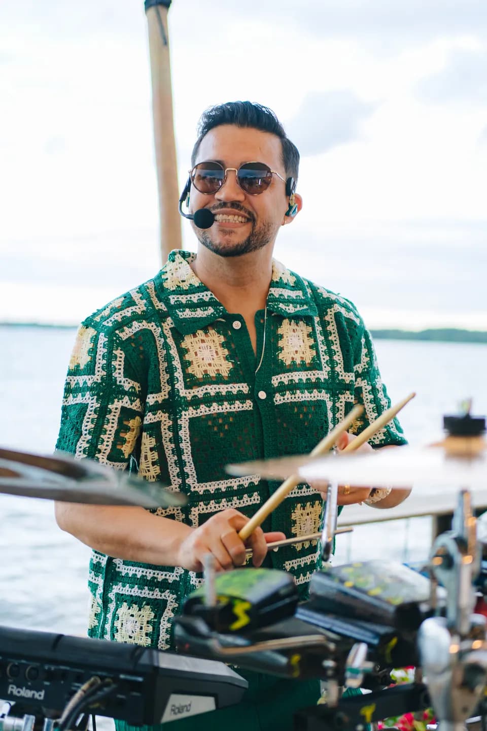 Saulo Torquato sorrindo com as baquetas na mão, atrás dos pratos, de camisa de crochê verde e branca